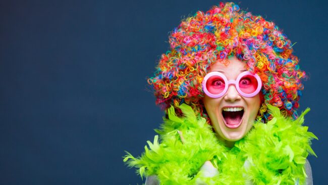 Person mit bunter Lockenperücke, pinker Brille und grüner Federboa vor dunkelblauem Hintergrund.