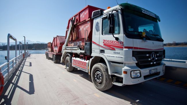 Ein Container-Lkw von Zosseder mit zwei Absetzcontainern steht auf einer Autofähre im Alpenvorland. Zosseder nutzt künftig eine digitale Portallösung zur effizienten Abwicklung von Kundenprozessen.