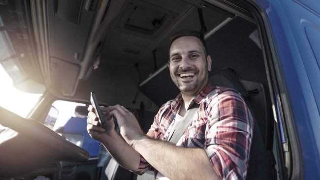 movatix-in-der-cloud Lkw-Fahrer lächelt und bedient ein Smartphone in der Fahrerkabine.