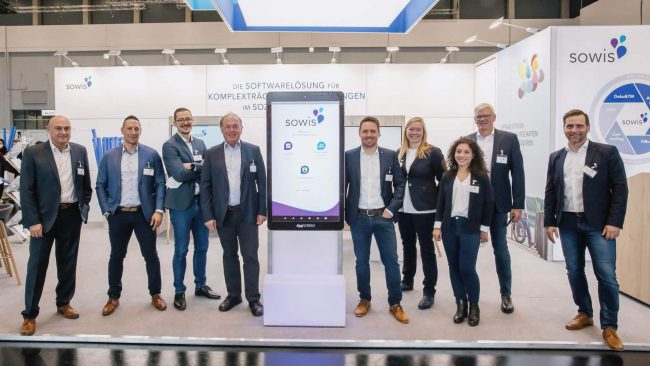 sowis-consozial-2021 Gruppenbild des sowis-Teams auf der ConSozial 2021 vor dem eigenen Messestand mit zentralem Touchdisplay und Präsentation der Softwarelösung.