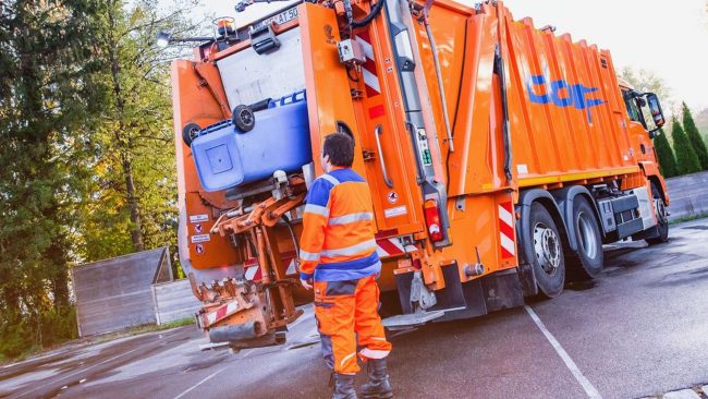 Dorr-Unternehmensgruppe-aus-Kempten-im-Allgäu-setzt-auf-das-Know-how-von-tegos Ein Müllwerker in orange-blauer Warnkleidung steht hinter einem Müllfahrzeug und beobachtet den automatisierten Entleerungsvorgang einer blauen Tonne. Die Dorr Unternehmensgruppe aus Kempten im Allgäu setzt auf das ERP-System enwis zur Digitalisierung ihrer Prozesse.