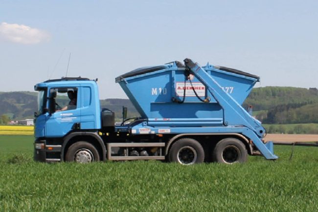 movatixPaperlessPickup-bei-der-Menshen-GmbH Blauer Lkw mit Absetzcontainer der A. Menshen GmbH & Co. KG auf einem Feld vor hügeliger Landschaft.