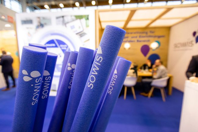Die-sowis-GmbH-nimmt-an-der-ConSozial-2023-teil—Vorreiter-für-Innovationen-in-der-Sozialwirtschaft Lila Werbestäbe mit sowis-Logo im Vordergrund, im Hintergrund Messestand und Gespräche auf der ConSozial 2023.