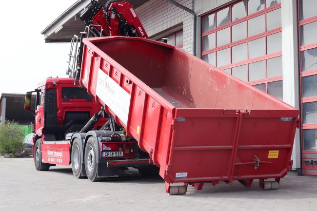 Hans-Meyer-aus-Erlangen-setzt-auf-enwis Abrollkipper der Firma Hans Meyer Entsorgungs GmbH aus Erlangen beim Absetzen eines roten Containers vor einer Halle.