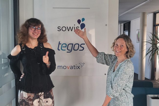 Neue-Auszubildende-bei-der-tegos-Holding-GmbH-Tabea-Breuer-startet-ihre-Reise-zur-Anwendungsentwicklerin Tabea Breuer, angehende Anwendungsentwicklerin, und ihre Ausbilderin vor einem Schild mit den Logos von sowis, tegos und movatix.