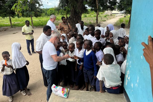 TEGOS-Holding-unterstützt-die-Kirschner-Stiftung-im-Kampf-für-Bildung-in-Bayern-und-Tansania Männer verteilen Luftballons an Schulkinder in Uniformen vor einem Schulgebäude in Tansania.