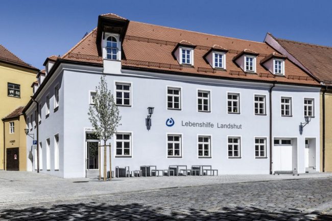 sowisDOKU-Lebenshilfe-Landshut_11-203 Außenansicht des Gebäudes der Lebenshilfe Landshut mit weißer Fassade und rotem Dach.