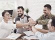 Lachendes Team legt im Büro motiviert die Hände übereinander, Symbol für Zusammenhalt und Dank.