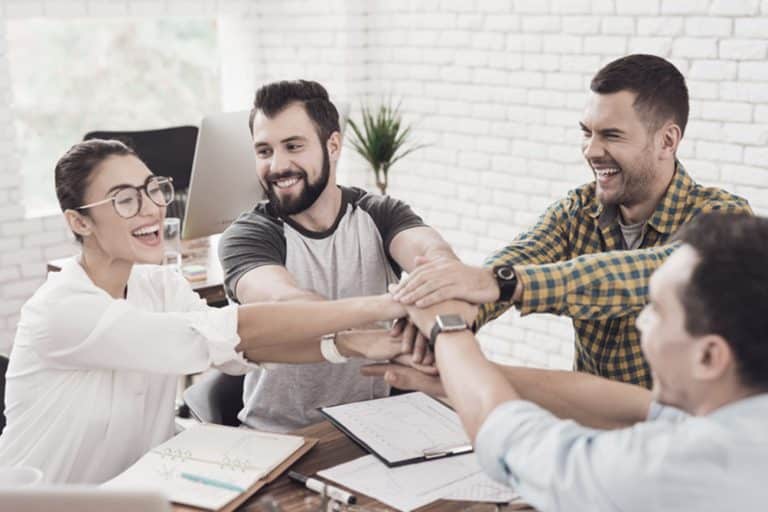Lachendes Team legt im Büro motiviert die Hände übereinander, Symbol für Zusammenhalt und Dank.