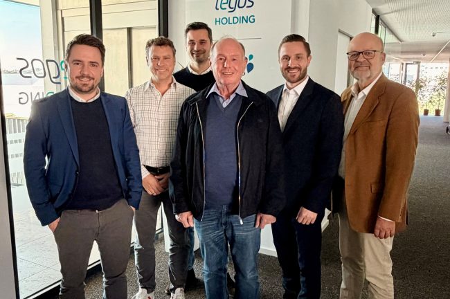 Siebte Beiratssitzung der tegos Holding. Daniel Artmann, Doktor Walter, Arthur Krautbauer, Professor Doktor B Niestroj, Maximilian Krautbauer und Michael Liebermann stehen vor einer Glaswand mit tegos Holding Logo nach einer erfolgreichen, gemeinsamen Sitzung.