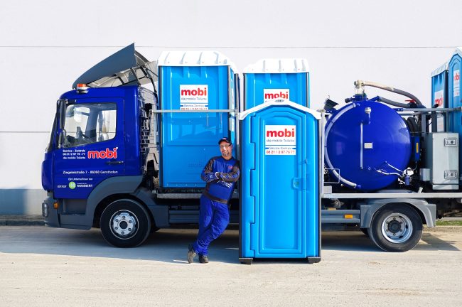 Ein Mann lehnt sich an eine mobile mobi Toilette vor einem Fahrzeug.