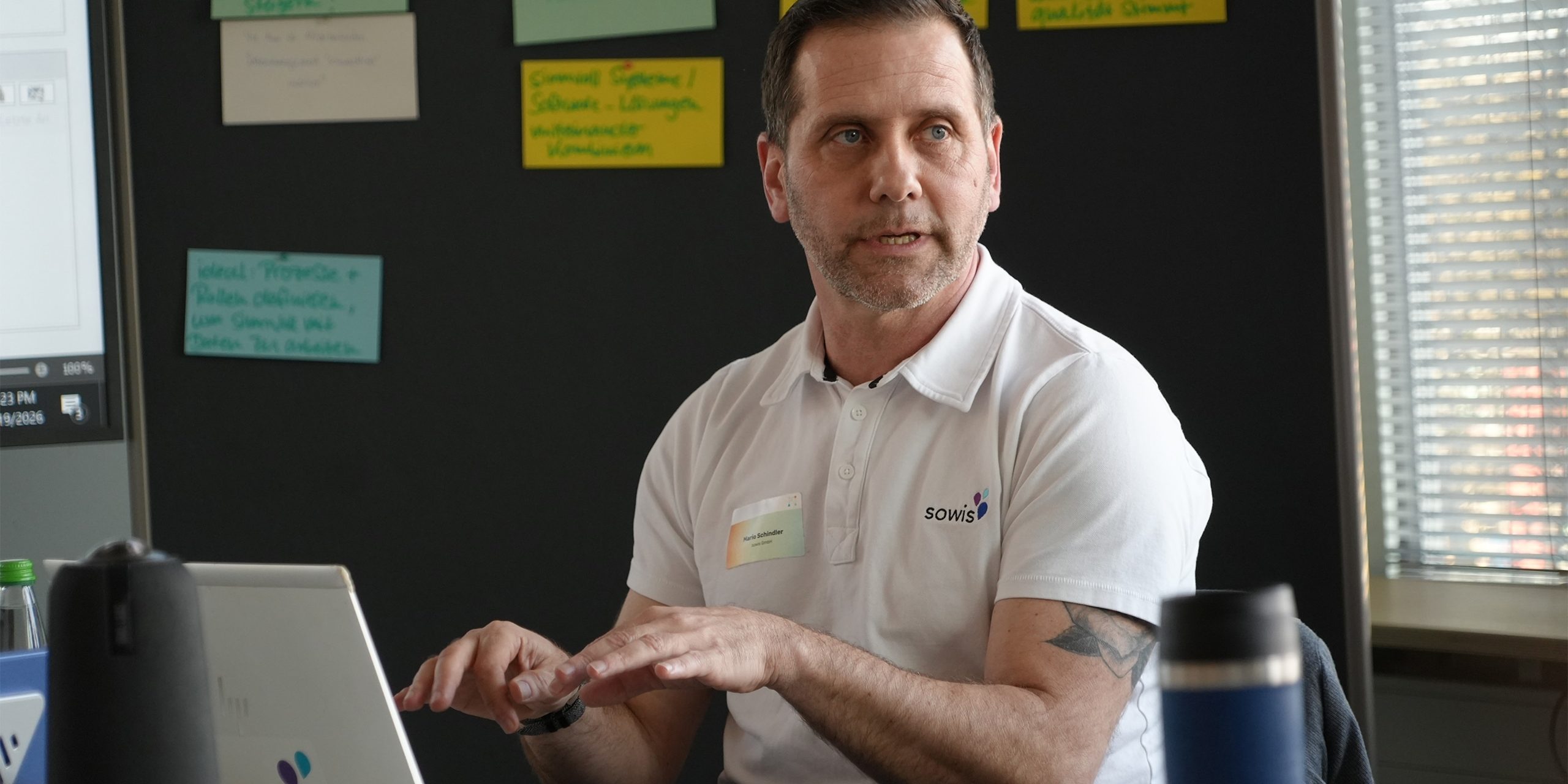 Ein Mann tipp auf seinem Laptop vor grauer Wand mit Notizen.